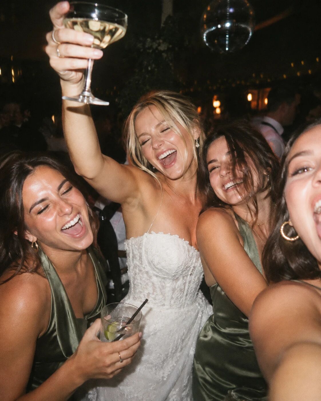 A bride laughing on the dancefloor, glass raised, surrounded by bridesmaids